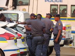 A group of South African Police Service (SAPS) officers standing next to patrol vehicles, wearing navy blue uniforms with SAPS badges and reflective vests, discussing operations in a city environment